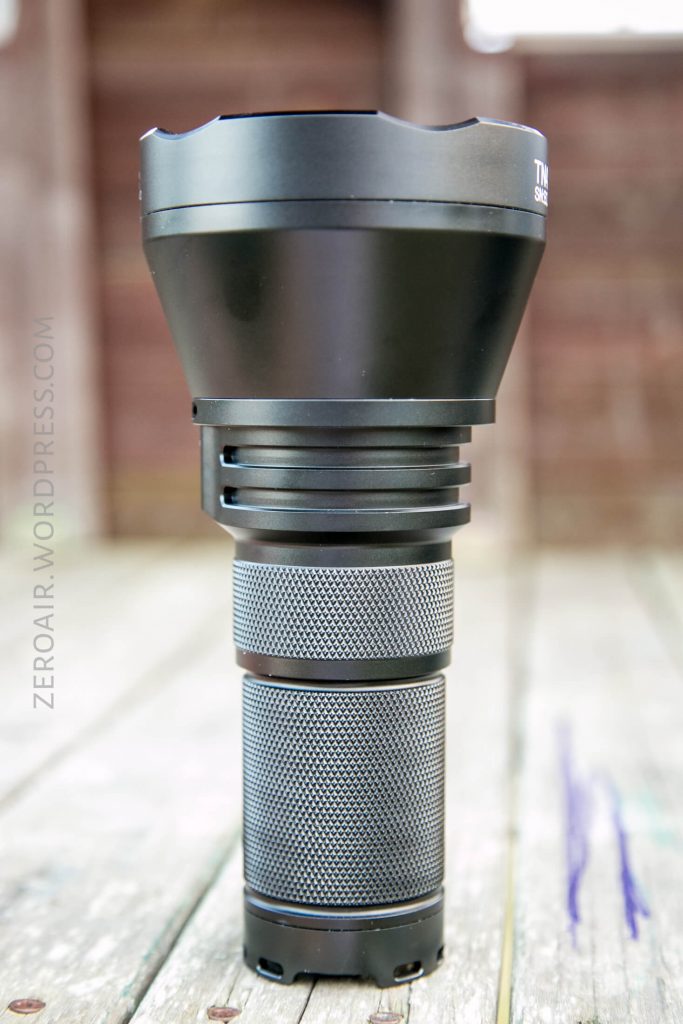 A large, black handheld flashlight with a textured grip stands upright on a weathered wooden surface, set against an out-of-focus wooden background.