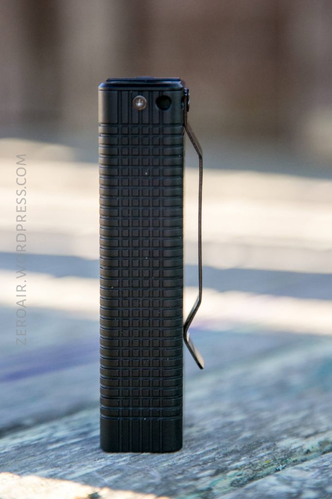 A black, rectangular flashlight with a textured grid pattern and a metal pocket clip is standing upright on a wooden surface. The background is blurred.