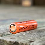 An orange Rofis 18650 rechargeable lithium-ion battery with 5000mAh capacity lies on its side on a weathered wooden surface.