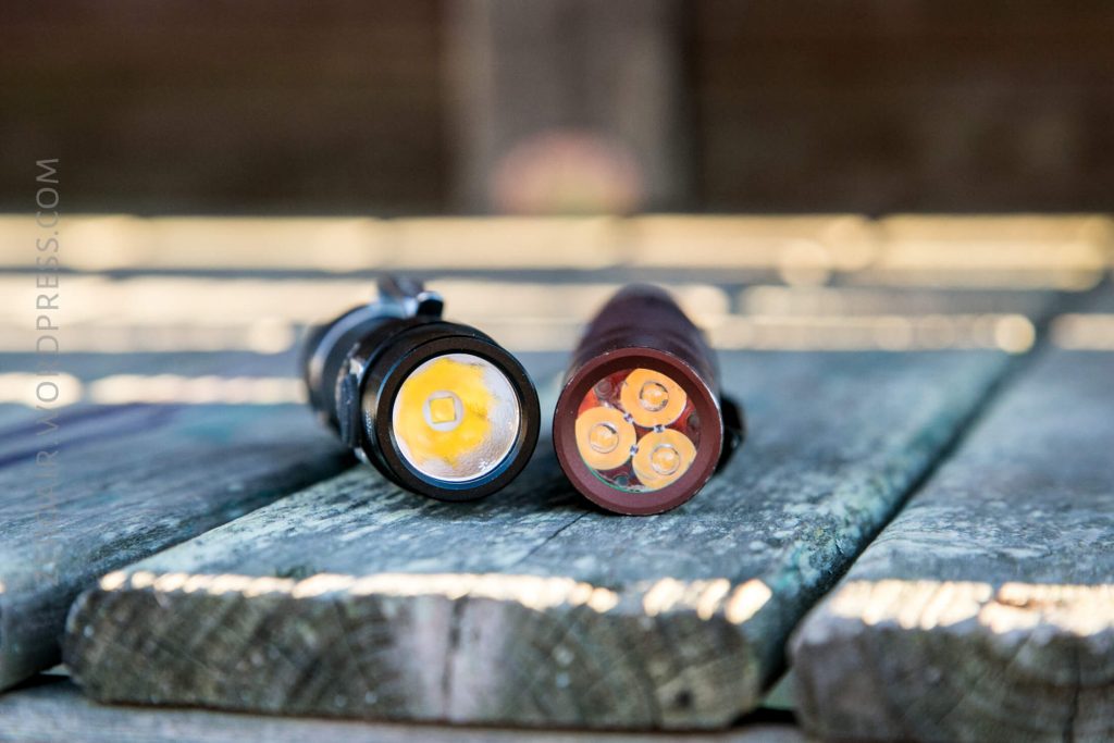 A close-up view of two flashlights lying on a wooden surface, positioned side by side with their lenses facing the camera. The left flashlight is black and the right one is brownish-red.