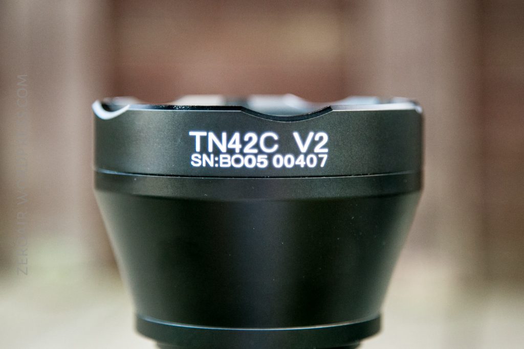 A close-up of a black cylindrical device labeled TN42C V2 SN: B005 00407 in white text, with a blurred brown background.
