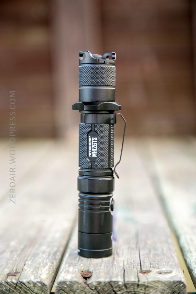 A black tactical flashlight stands upright on a weathered wooden surface, with a metal clip on its side and textured grip sections. The background is blurred.