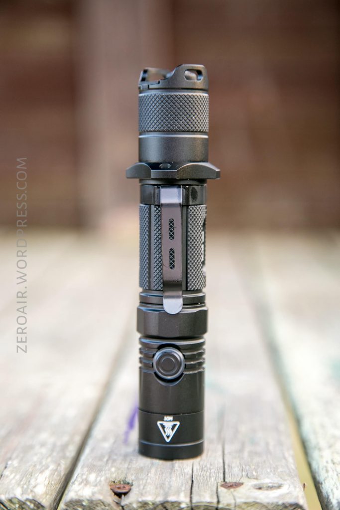 A black tactical flashlight is standing upright on a weathered wooden surface. The background is blurred and the flashlight features a textured grip and a side button. The text “ZEROAIR.WORDPRESS.COM” is visible on the left side.