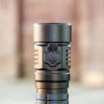 A close-up of a black flashlight standing upright outdoors, with the word OPEN and an arrow engraved on a latch near the head of the flashlight. The background is blurred.