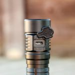 A close-up of a flashlight with a rubber cover open to reveal a micro USB charging port on its side. The flashlight has a metallic finish and a grooved design. The background is blurred.