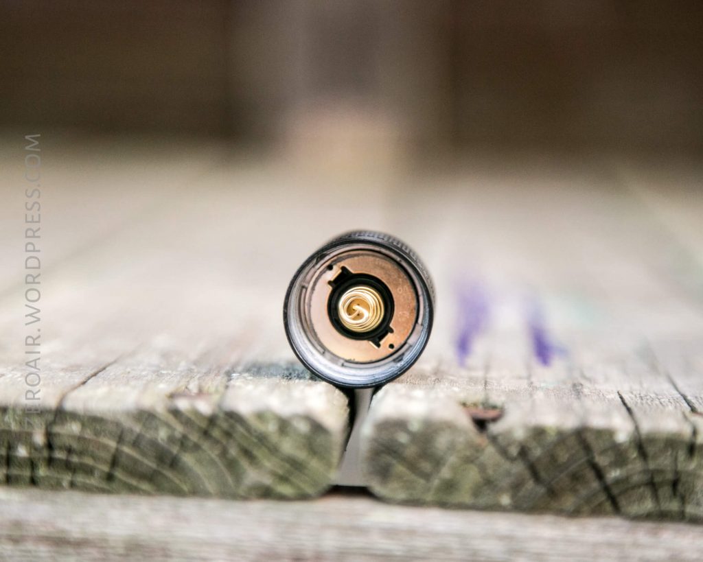 Close-up view of the bottom end of a cylindrical metal object, possibly a flashlight or battery, resting on weathered wooden boards with a blurred background.