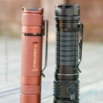 Two flashlights standing upright on a wooden surface. The left one is copper-colored with a label reading Convoy, and the right one is black and larger with a more textured body. Both have metal pocket clips.