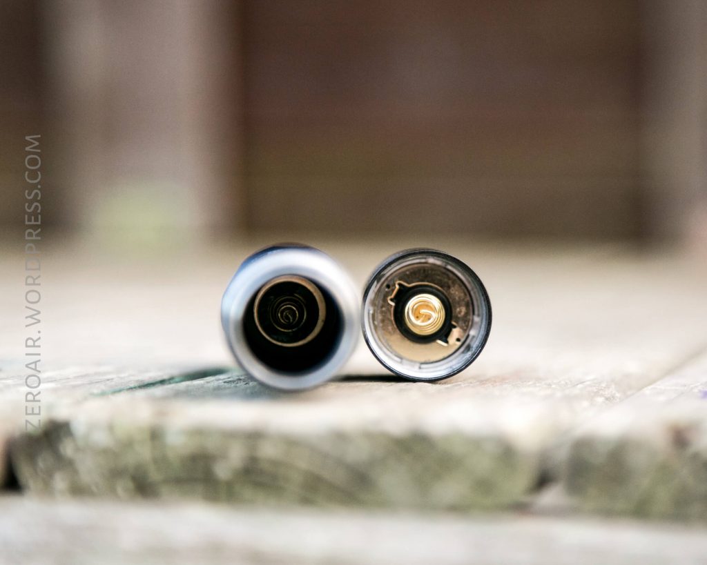 Close-up of two cylindrical metal objects, possibly flashlight battery compartments, placed horizontally on a wooden surface with their open ends facing the camera, showing coiled metal springs inside.