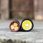 Close-up of two cylindrical flashlight heads lying side by side on a wooden surface, showing their LED emitters and textured reflectors; one has three LEDs, the other has a single LED.