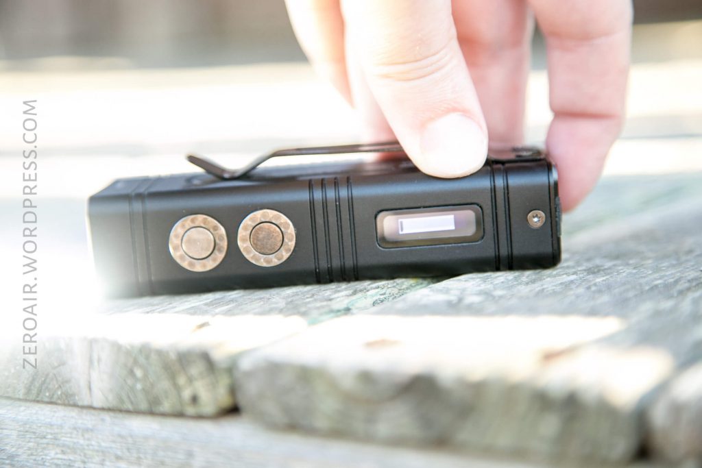A hand picks up a small black rectangular device with two circular buttons and a clip, resting on a wooden surface. The device has a small display screen. The website zeroair.wordpress.com is visible on the left.