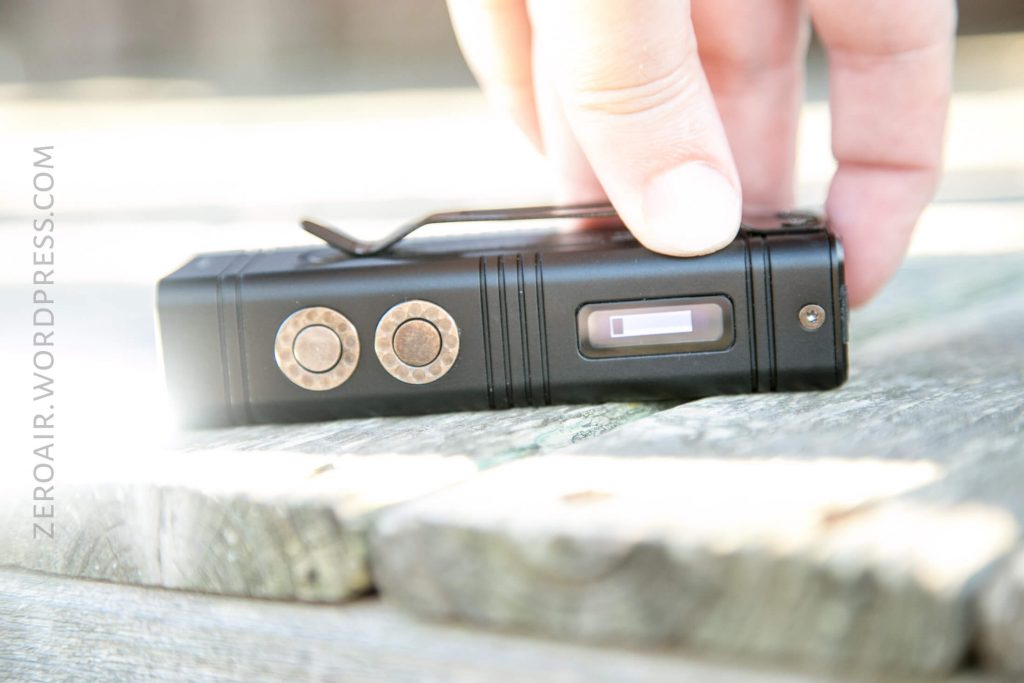 A close-up of a person’s hand holding a small black electronic device with two round buttons, a belt clip, and a small display screen, placed on a wooden surface.