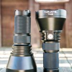 Two black handheld flashlights, labeled “ThruNite TN42” and “TN42 V2,” stand upright on a wooden surface outdoors. The flashlights have textured grips and large lens heads. A blurred building is in the background.