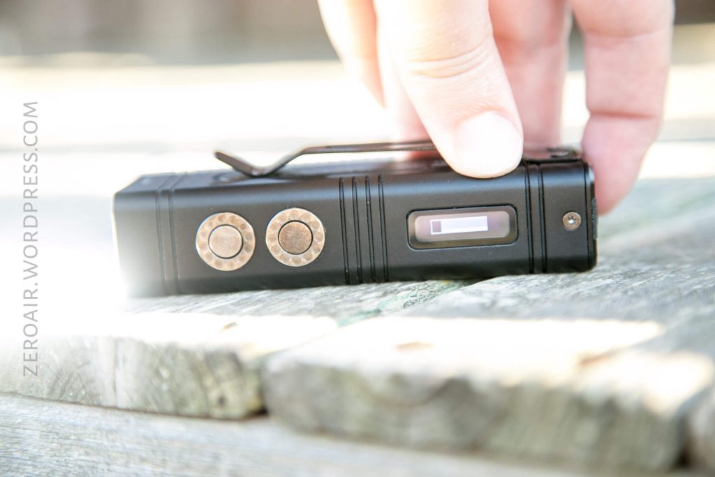 A hand holds a small black electronic device with two round metal buttons and a small rectangular screen, resting on a wooden surface. The words ZEROAIR.WORDPRESS.COM are visible along the left edge.