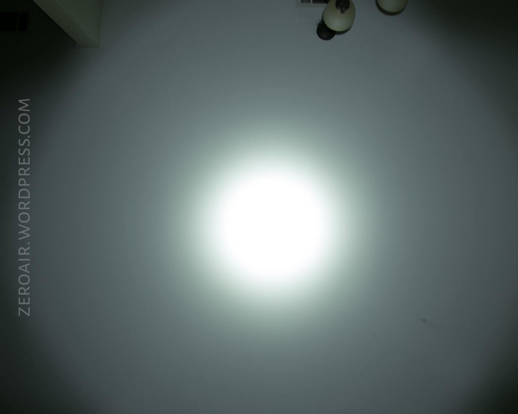 A bright white flashlight beam creates a circular spot on a light-colored wall. Two ceiling light fixtures are partially visible at the top right. ZEROAIR.WORDPRESS.COM is written vertically along the left side.