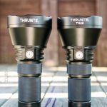 Two black handheld flashlights labeled ThruNite are standing upright side by side on a wooden surface. The flashlight on the right is labeled TN42. Sunlight shines on the flashlights and the wood.