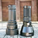 Two large, black flashlights stand upright side by side on a wooden surface outdoors. Sunlight highlights their metal bodies. The left one is bulkier, while the right one has THRUNITE branding on its base.