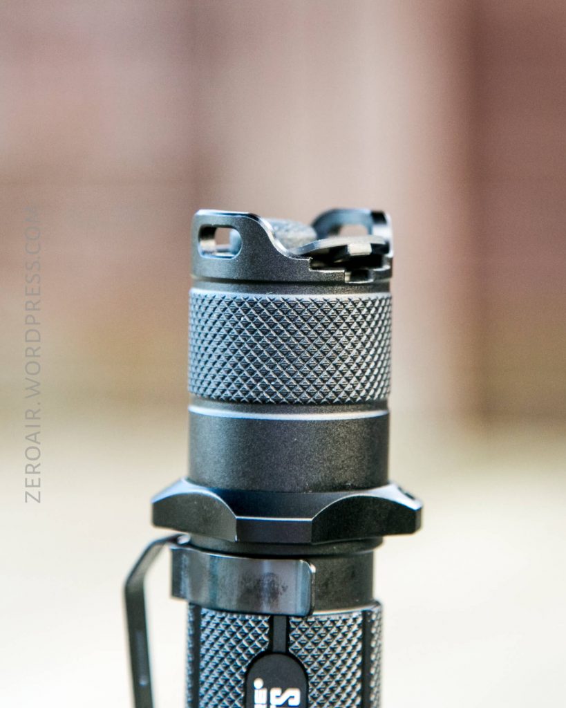 A close-up view of the textured grip and tail cap of a black flashlight, displaying its knurled surface and a lanyard attachment point, with a blurred background.