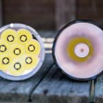 Two flashlight heads are shown close-up on a wooden surface. The left flashlight has seven LED emitters arranged in a hexagonal pattern, while the right flashlight has a single centered LED emitter.