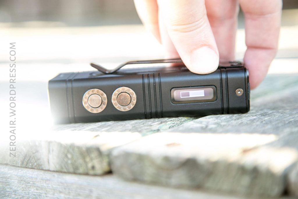 A close-up of a person’s hand reaching for a small black rectangular electronic device with circular metal buttons and a digital display, placed on a wooden surface. The text ZEROAIR.WORDPRESS.COM is visible on the left side.
