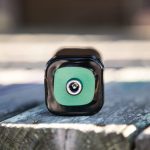 A close-up of a black rectangular battery lying on its side on a wooden surface, with the positive terminal and a green label facing the camera.