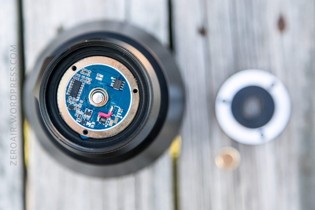 Close-up of an electronic circuit board inside a cylindrical device, with a detached circular cover and a small round metal piece placed on a wooden surface nearby.