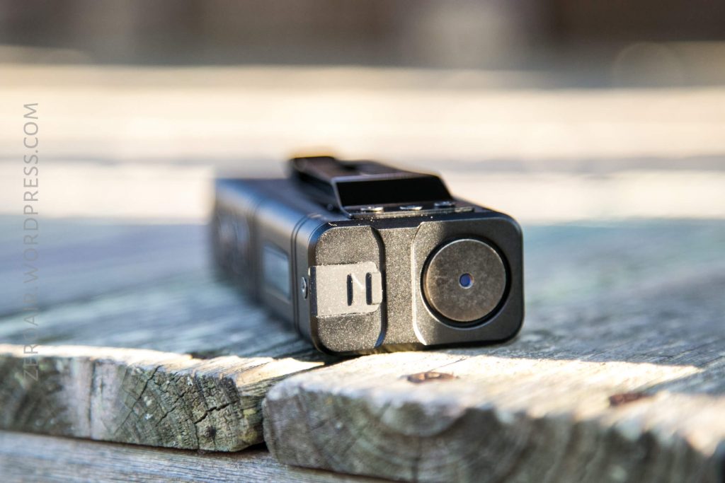A compact black body camera rests on a weathered wooden surface, viewed from the front with a lens and button visible. Sunlight creates bright highlights and shadows.