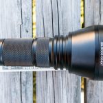 A black flashlight labeled TN42C V2 SN:B5050407 lies horizontally on a weathered wooden surface next to a faintly visible printed card. ZEROAIR.WORDPRESS.COM is written vertically on the left side.