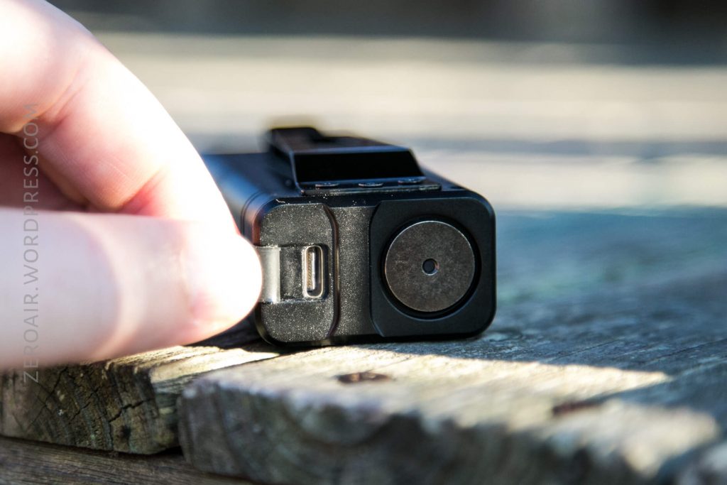 A close-up of a hand holding a black electronic device with a USB-C port and a round magnetic or metallic contact, resting on a wooden surface outdoors.