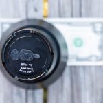 A cylindrical object labeled BP10 V2 is positioned vertically on a wooden surface, with a U.S. one-dollar bill placed flat underneath it. The website ZEROAIR.WORDPRESS.COM is visible along the left side.