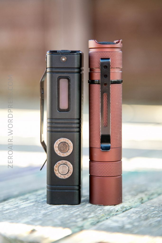 Two flashlights standing upright on a wooden surface. The left flashlight is black with two buttons and a rectangular window, while the right flashlight is reddish-brown with a textured grip and a black clip.