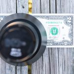 A metal combination safe lock covers part of a two-dollar bill lying on a weathered wooden surface. The bill is partially visible, with the serial number and seal clearly shown.