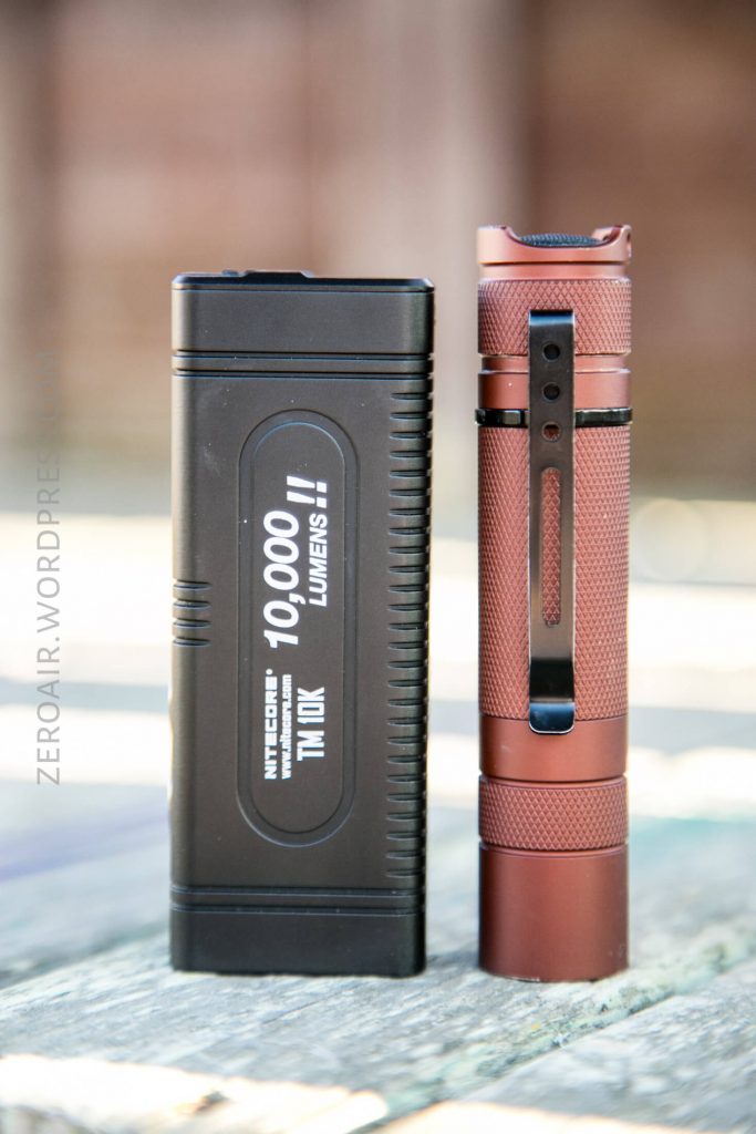 Two flashlights are standing upright on a wooden surface. The left flashlight is black and rectangular with “10,000 lumens!!” printed on the front, and the right flashlight is cylindrical and copper-colored with a pocket clip.