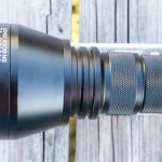 A close-up of a black flashlight labeled TN42C V2 SN:B005 00047 lying horizontally on a weathered wooden surface. The head and part of the textured grip are visible.