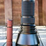 A red compact flashlight stands next to a large black flashlight labeled TN42C V2 SN: 80050407 on a wooden surface with a blurred wooden wall in the background.