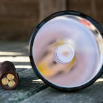 A large LED flashlight is lying on its side on a wooden surface, with the lens and bulb facing the camera. Next to it is a battery pack containing four visible cylindrical batteries.