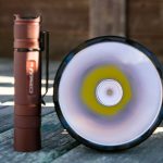 A brown flashlight stands upright next to a larger flashlight, which is positioned lens-up to show the wide reflector and LED, on a wooden surface with a blurred background.