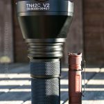 A large black TN42C V2 flashlight stands upright next to a much smaller brown flashlight with a black clip, both placed on a wooden surface outdoors with a blurred wooden fence in the background.
