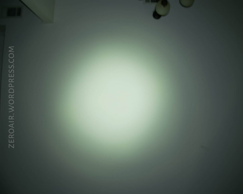 A flashlight casts a bright white circular beam on a wall in a dim room. Ceiling light fixtures and air vents are partially visible at the top. ZEROAIR.WORDPRESS.COM is written along the left side.