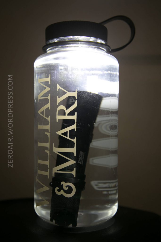 A clear plastic water bottle with WILLIAM & MARY written vertically in white letters, filled with water and containing a black flashlight inside. The bottle cap is black with a loop.