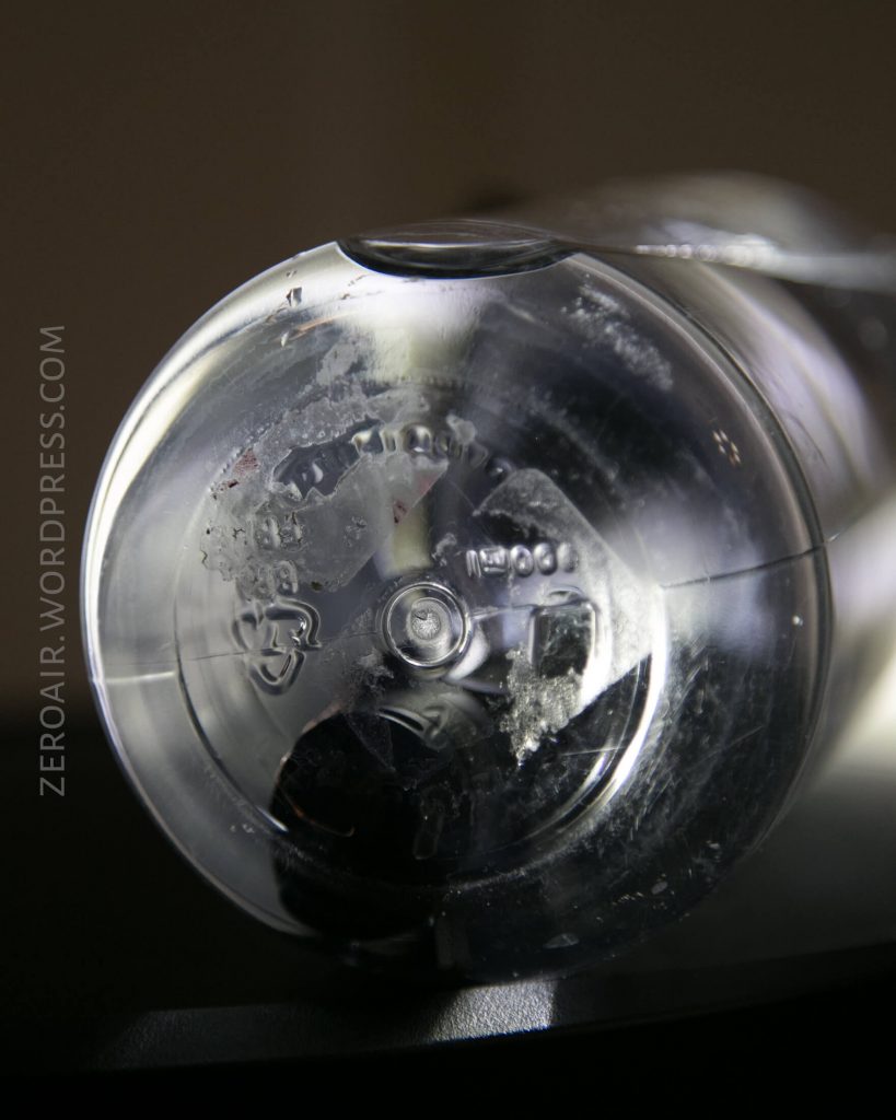 Close-up of the bottom of a transparent plastic bottle, showing embossed recycling symbols, numbers, and letters, with light reflecting off the surface. The text ZEROAIR.WORDPRESS.COM is visible along the left edge.