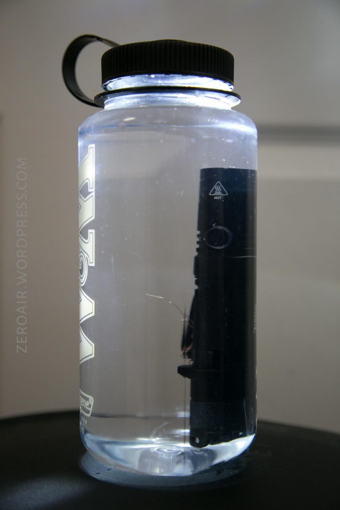 A flashlight is illuminated while submerged in a large, transparent water bottle with a black lid and handle, sitting on a dark surface. The bottle is filled with clear water.