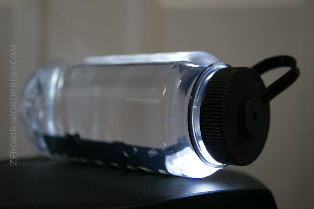 A clear plastic water bottle with a black screw cap lies on its side on a dark surface. The bottle is filled with water and illuminated from behind. A black strap is attached to the cap.