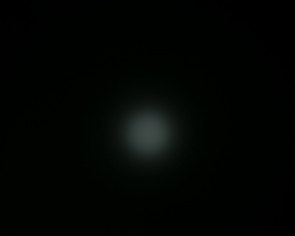 A faint, circular, white light glows in the center of a dark, nearly black background. The light appears slightly blurred and diffused, with no discernible features or objects surrounding it.