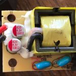 Close-up of an electronic circuit board showing capacitors labeled 820 6.3V SS812, a yellow-wrapped transformer, wires, blue components, and white adhesive securing parts to the board.