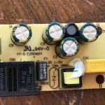 A close-up of an electronic circuit board with four large capacitors, a black relay, yellow fuse, several resistors, labels, and component markings visible on a wooden surface background.