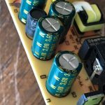 Close-up of an electronic circuit board featuring four blue-green cylindrical capacitors with yellow labels, a black cylindrical component, a yellow rectangular component, and visible circuit traces.