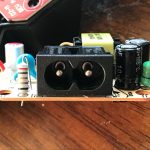 A close-up view of an electronic circuit board featuring a black two-prong power connector, capacitors, resistors, and various components, set on a wooden surface.
