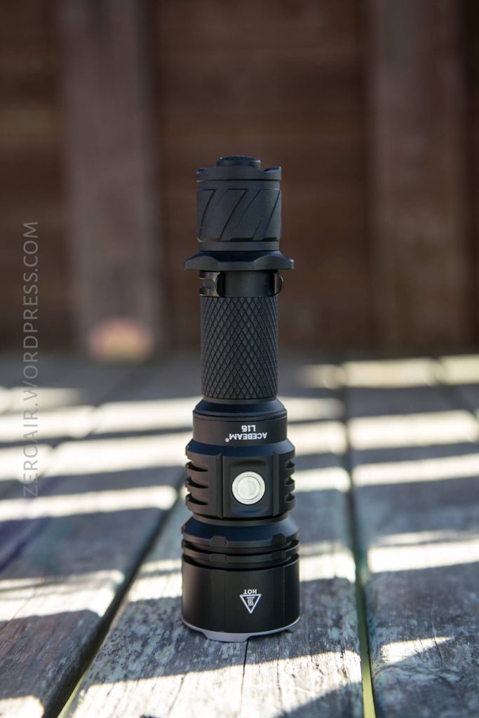 A black Acebeam L18 tactical flashlight stands upright on a wooden surface with sunlight casting shadows. The background is blurred, emphasizing the flashlight’s textured grip and prominent power button—perfect alongside the Acebeam L16 Flashlight.