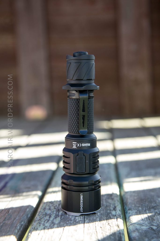 An Acebeam L16 Flashlight stands upright on a wooden surface, featuring a textured grip and several ridges along the body. The background consists of blurred wooden panels.