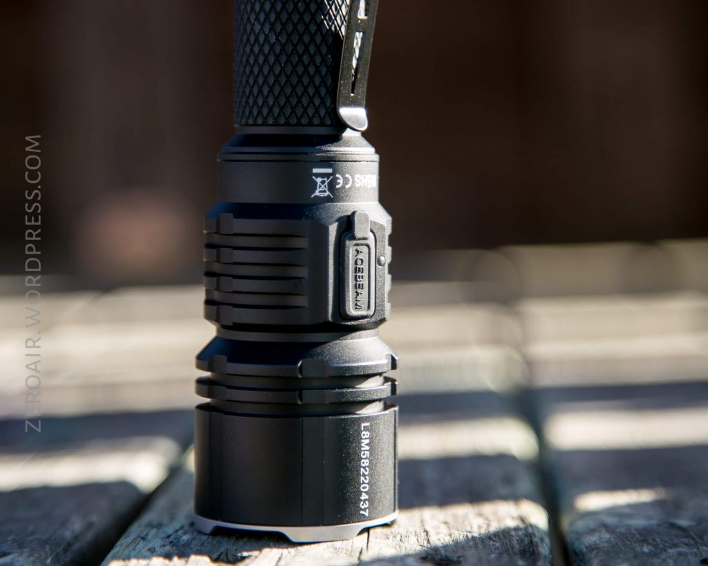 A close-up of an Acebeam L16 Flashlight standing upright on a wooden surface, with clear views of its textured grip, a side button, and serial number markings. The background is blurred.